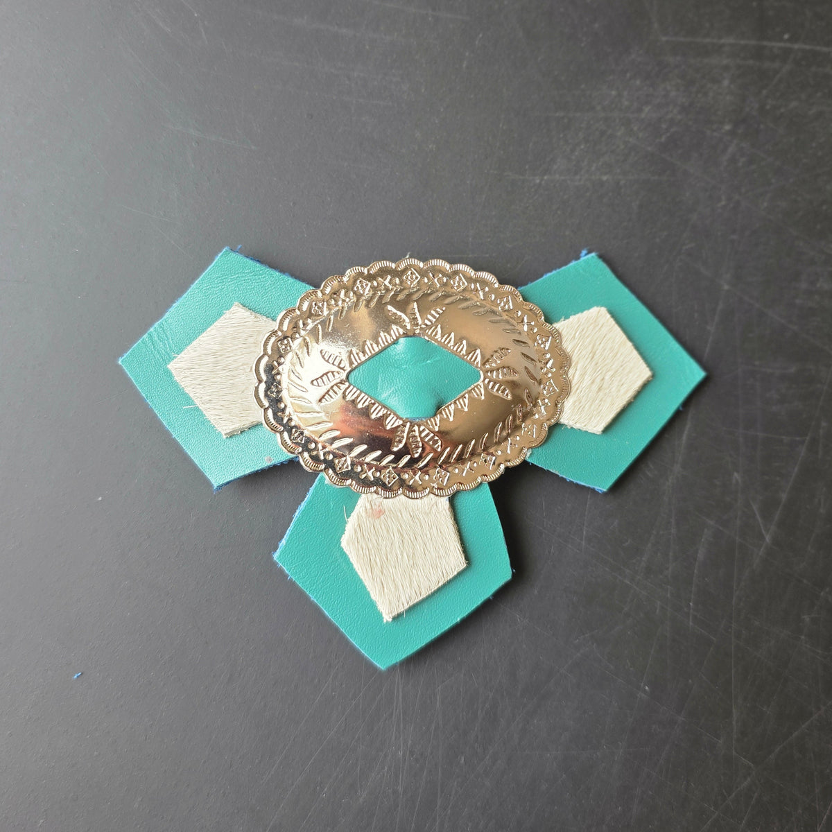 A turquoise and cream leather accessory with a central gold-toned concho buckle, photographed on a dark gray textured surface.